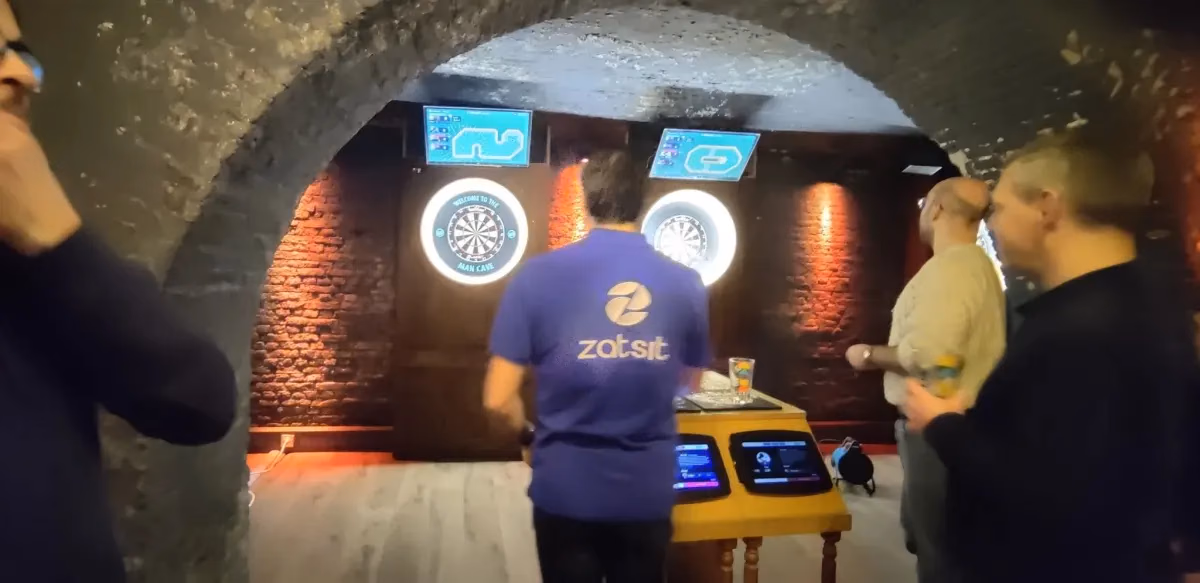 Partie de fléchettes dans une cave voûtée en briques, un joueur de dos en polo zatsit face à la cible
