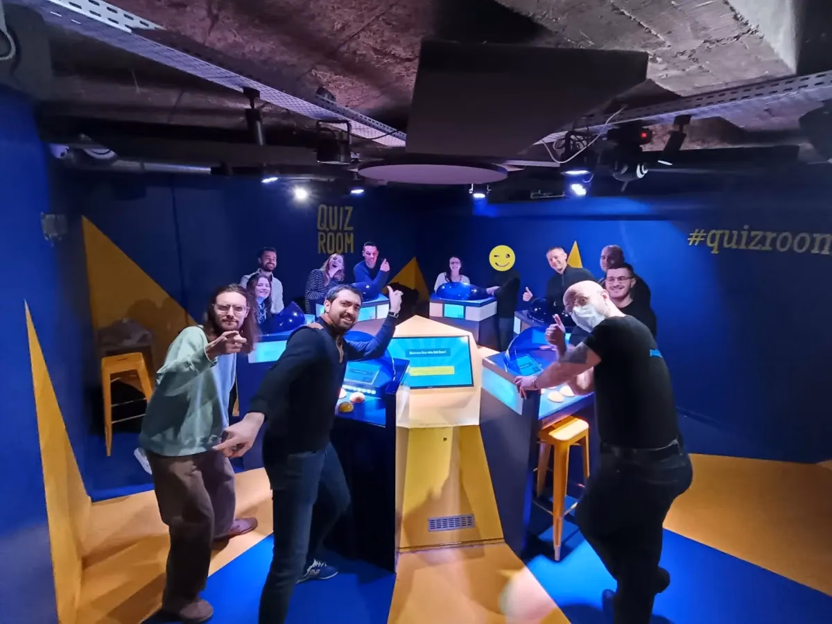 Le collectif dans une salle de Quiz Room aux murs bleus et jaunes, rassemblé autour des pupitres de jeu, pouces levés vers l'objectif